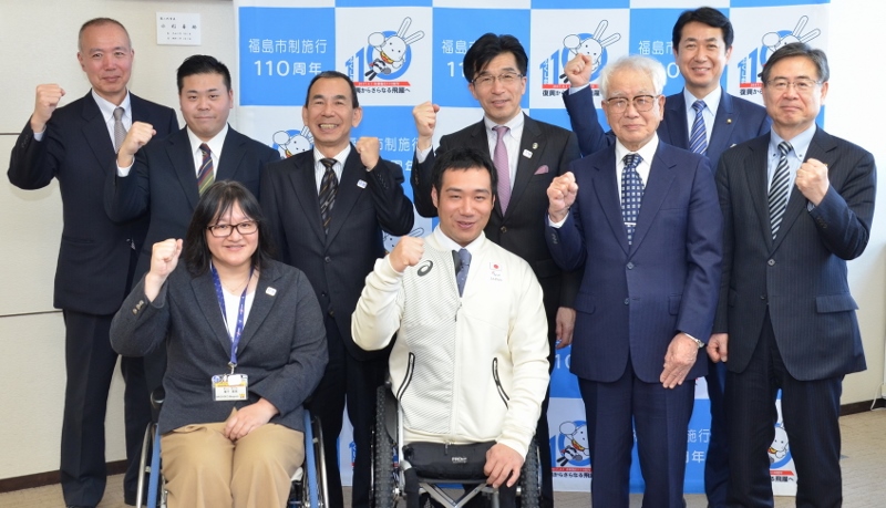 市長とアルペンスキー代表の鈴木猛史選手と関係者9名がバックパネルの前に並びガッツポーズを取っている写真