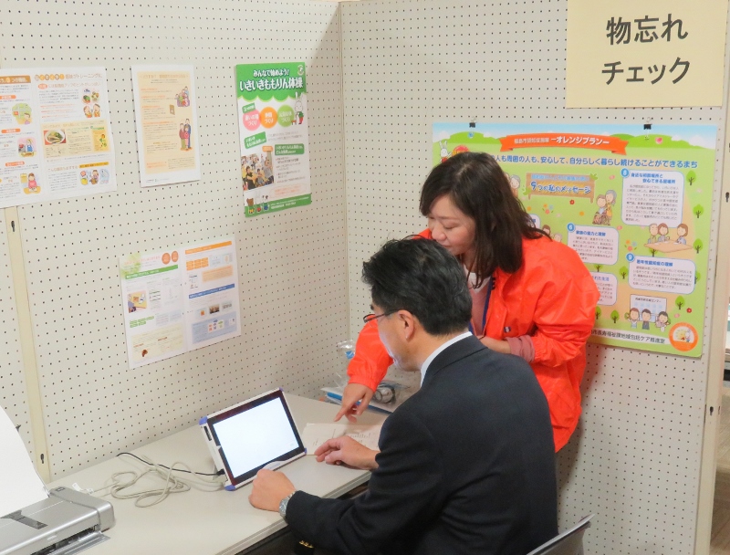 部屋の一室で市長が椅子に座り、関係者と一緒に液晶画面を見ている写真