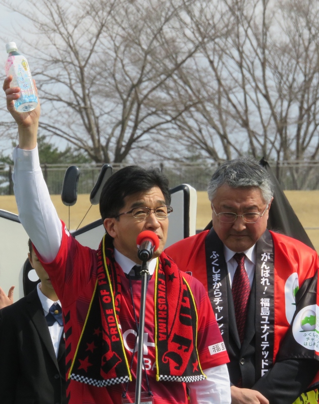 赤いユニフォーム姿の市長がタオルを首に掛け、右手にペットボトルを手に持ってマイクの前で話をしている写真