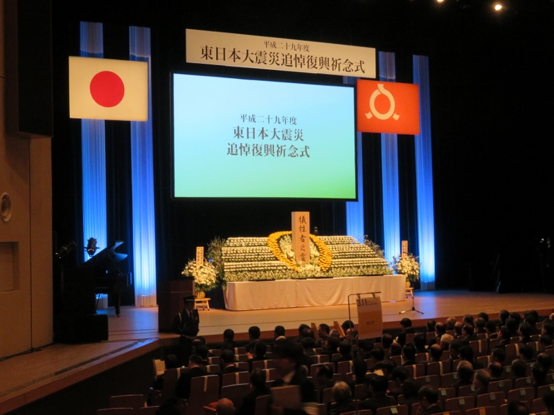 「東日本大震災追悼復興祈念式」のパネルが掛けられており、祭壇に花が飾られた式典の様子の写真