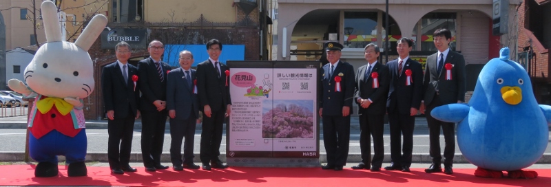 両端にももりんとマカプゥが手を広げてポーズを取っており、その間に市長と関係者が配電用地上機器を挟んで並んでいる写真