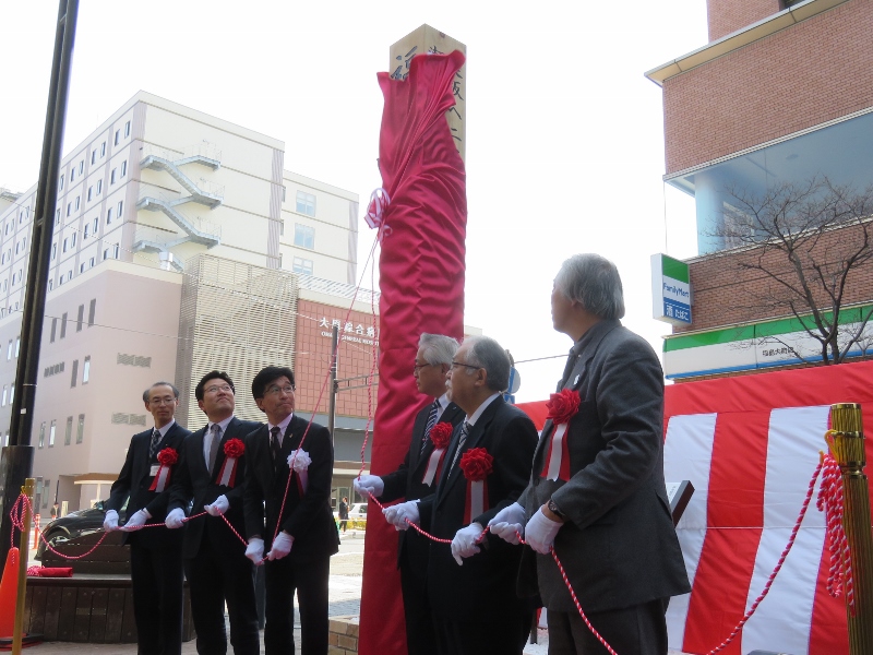 元標が赤い幕で覆いかぶせており、市長と関係者赤い紐を引きながら幕を取ろうとしている除幕式の様子の写真