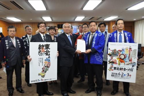スーツの男性と青や紺の法被を着た男性たちの前で、祭りの看板を持った男性たちの間に立った2人が書類を一緒に持っている写真