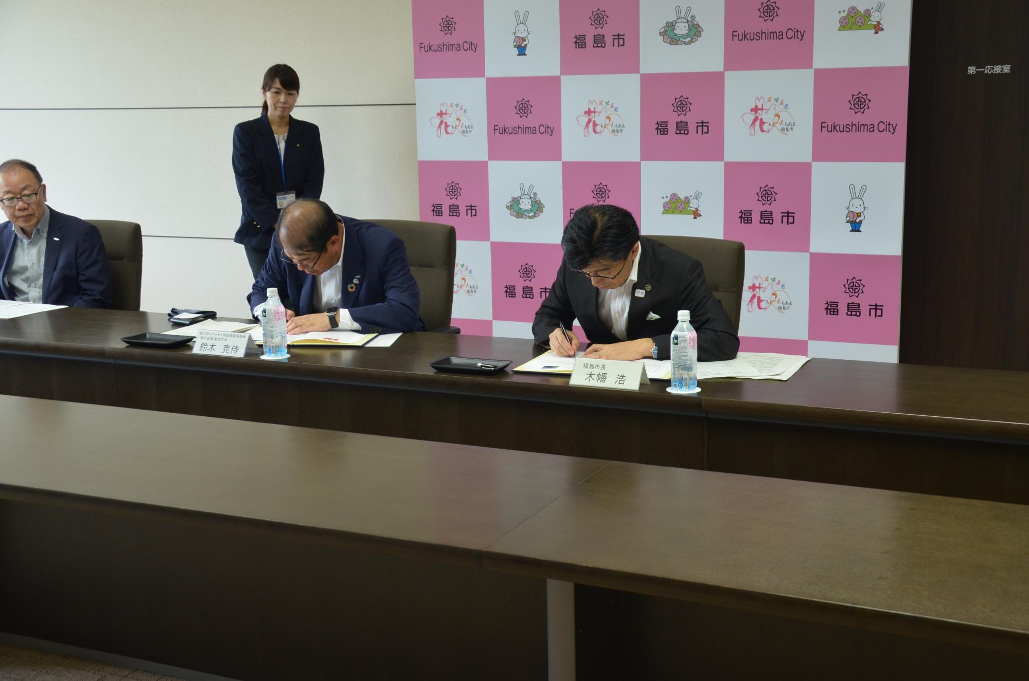 福島市のバックボードが置かれた壁を背に席に座った男性2名が机の書類に書き込んでいる様子の写真
