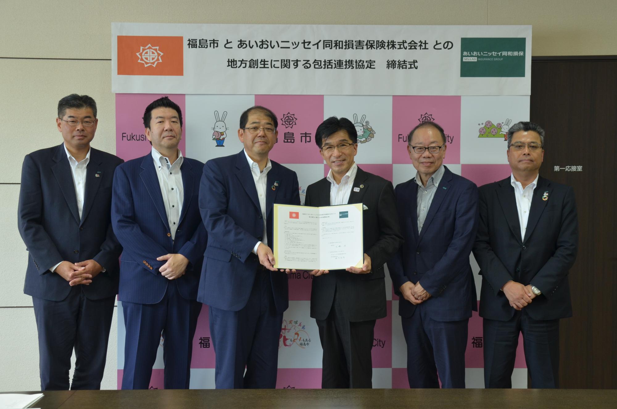 福島市のバックボードの前で、協定書を左右から持った市長と紺色のスーツ姿の男性を中央にして、他4名のスーツ姿の男性が並んで写った記念写真