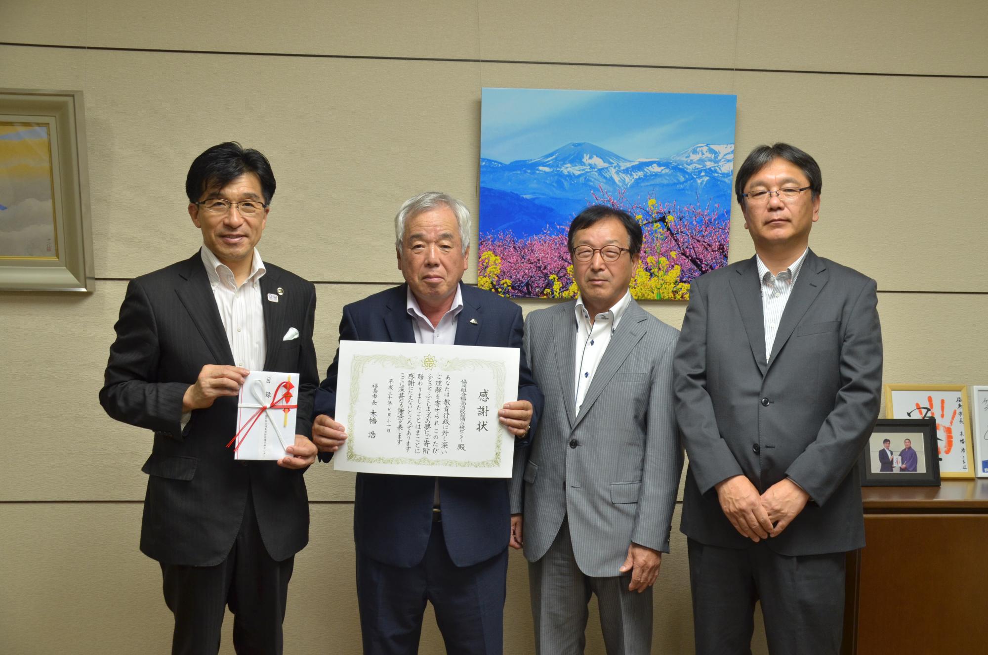 山の写真が飾られた壁の前で、寄付金の封筒を持った市長、賞状を持った男性他2名の関係者の方が並んで写った記念写真