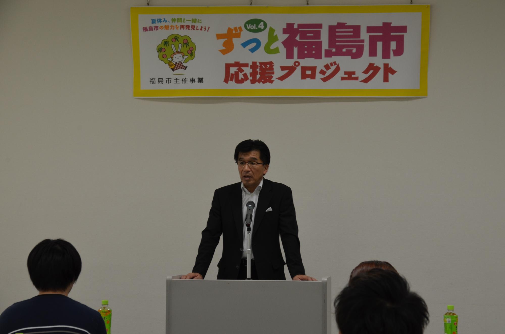 「ずっと福島市応援プロジェクト」のパネル前で演壇に立ち、話をする市長と耳を傾ける参加者の写真