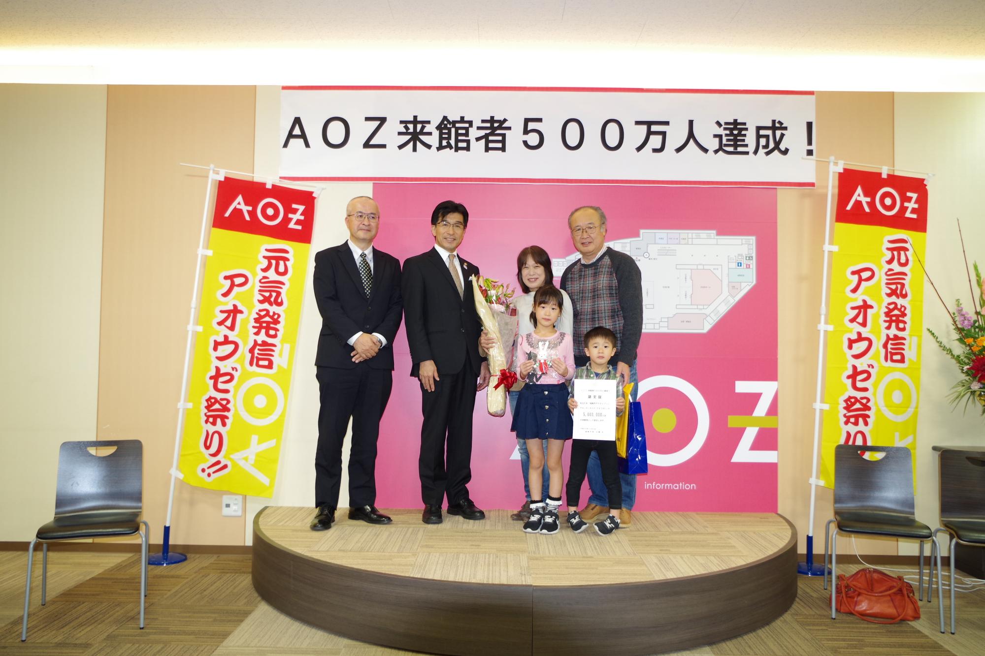 「AOZ来館者500万人達成!」のパネル前に立っている市長と花束を手に持った親子連れの写真