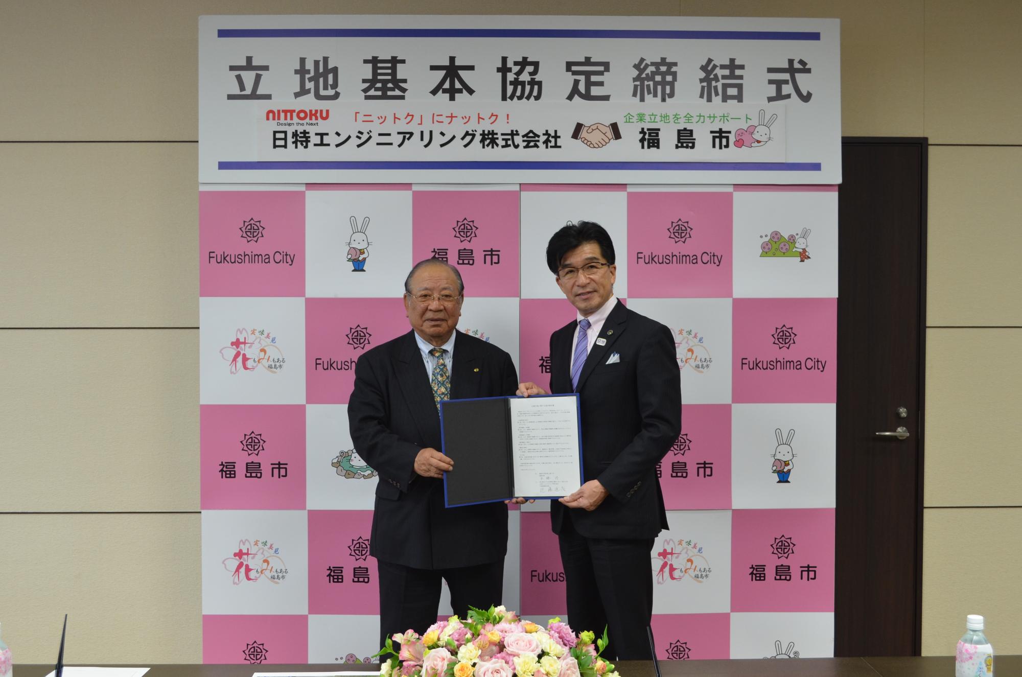 「立地基本協定締結式」のパネルが掲げられており、開いた締結書を手に持つ市長と近藤社長の写真