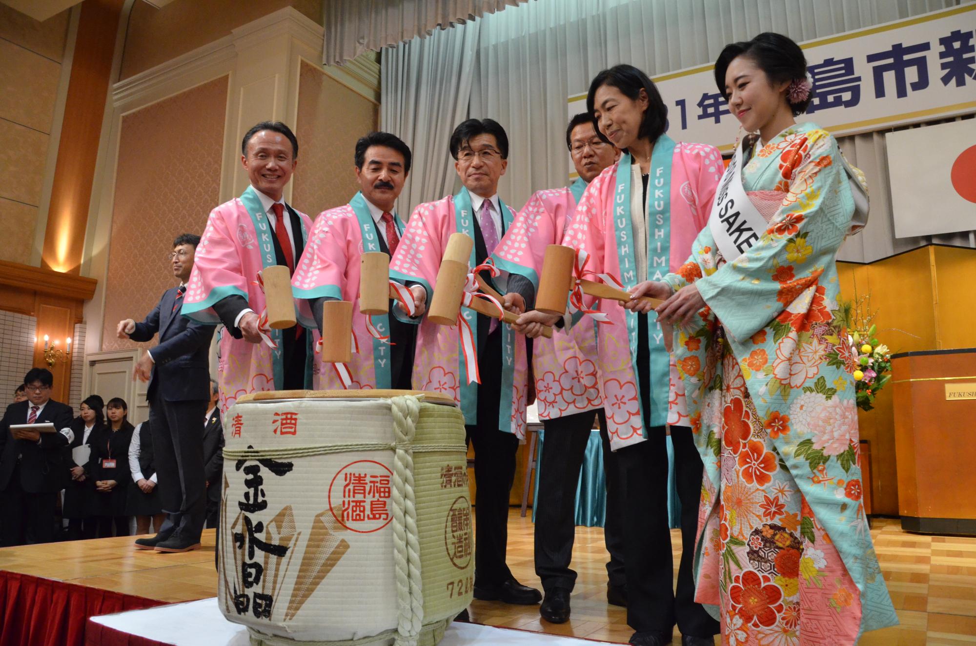 ステージの上で桜色のはっぴ姿で小槌を手に持つ市長と関係者5名が並び、鏡開きをしている写真