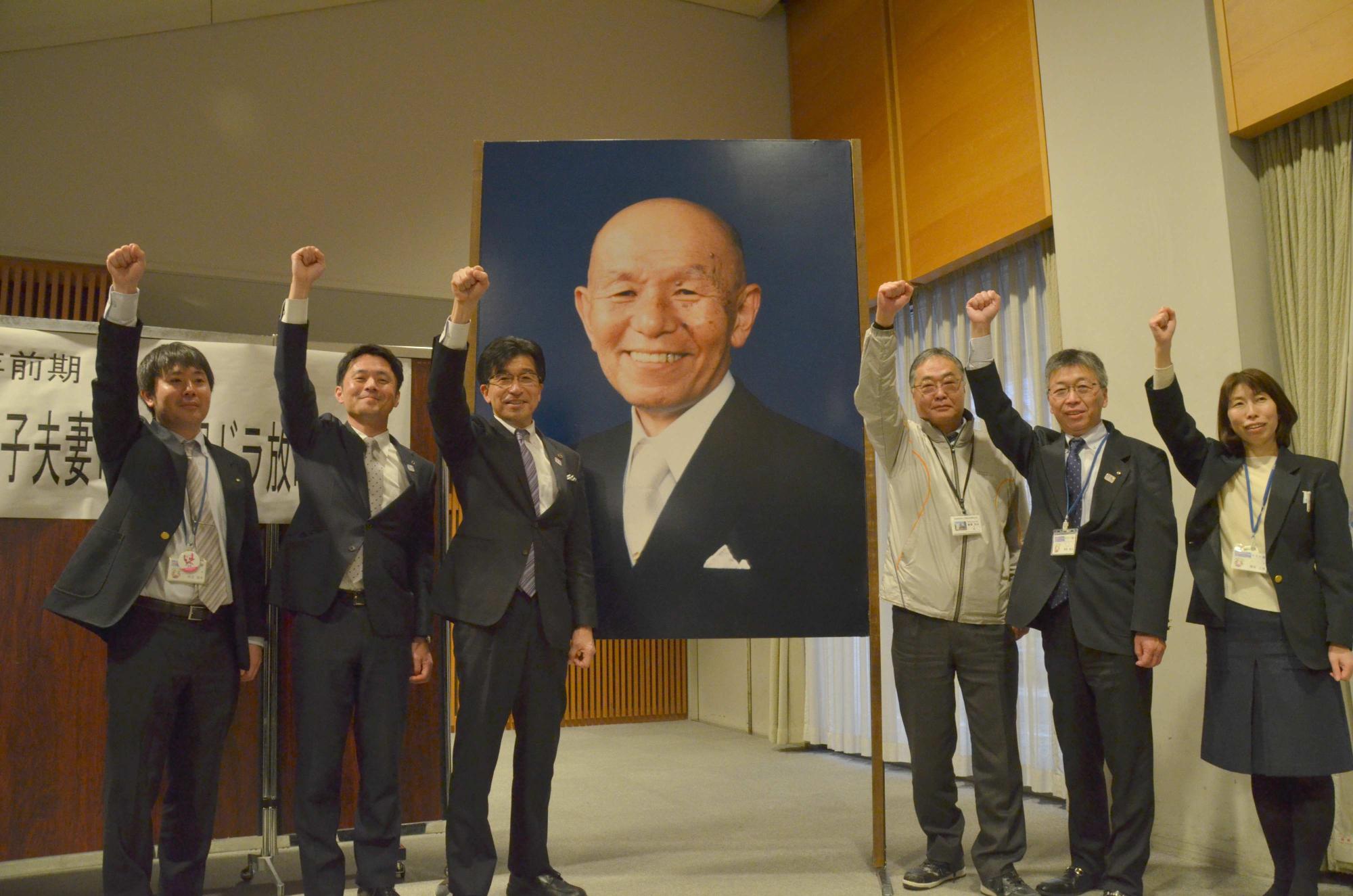 古関裕而さんの顔写真パネルのサイドで、市長と関係者5名が右手の拳を上げている写真
