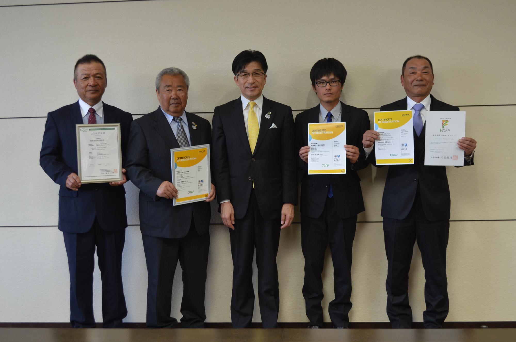 スーツを着た市長と両脇にGAP認証を両手に持った男性がそれぞれ2名ずつ横並びで立ち、記念撮影をしている写真