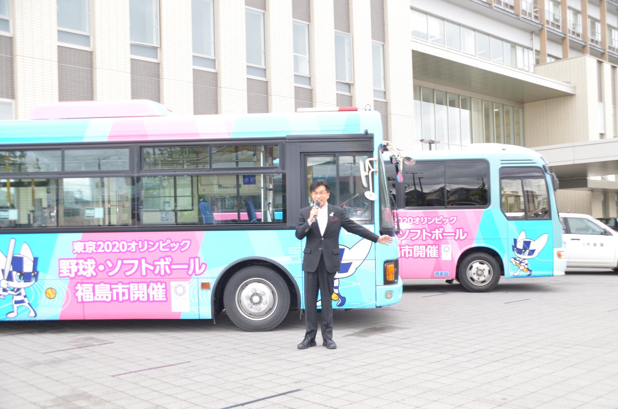 ピンク色と水色の車体に東京2020オリンピックマスコットキャラクターのミライトワがのったラッピングバスの前であいさつをする市長の写真
