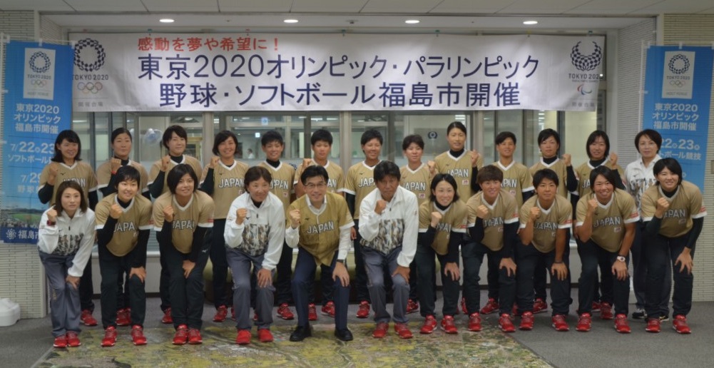 「感動を夢や希望に!東京2020オリンピック・パラリンピック野球・ソフトボール福島市開催」と書かれた横断幕の下で、ベージュのユニホームを着た市長とソフトボール女子日本代表の選手たちがガッツポーズをし記念撮影している写真
