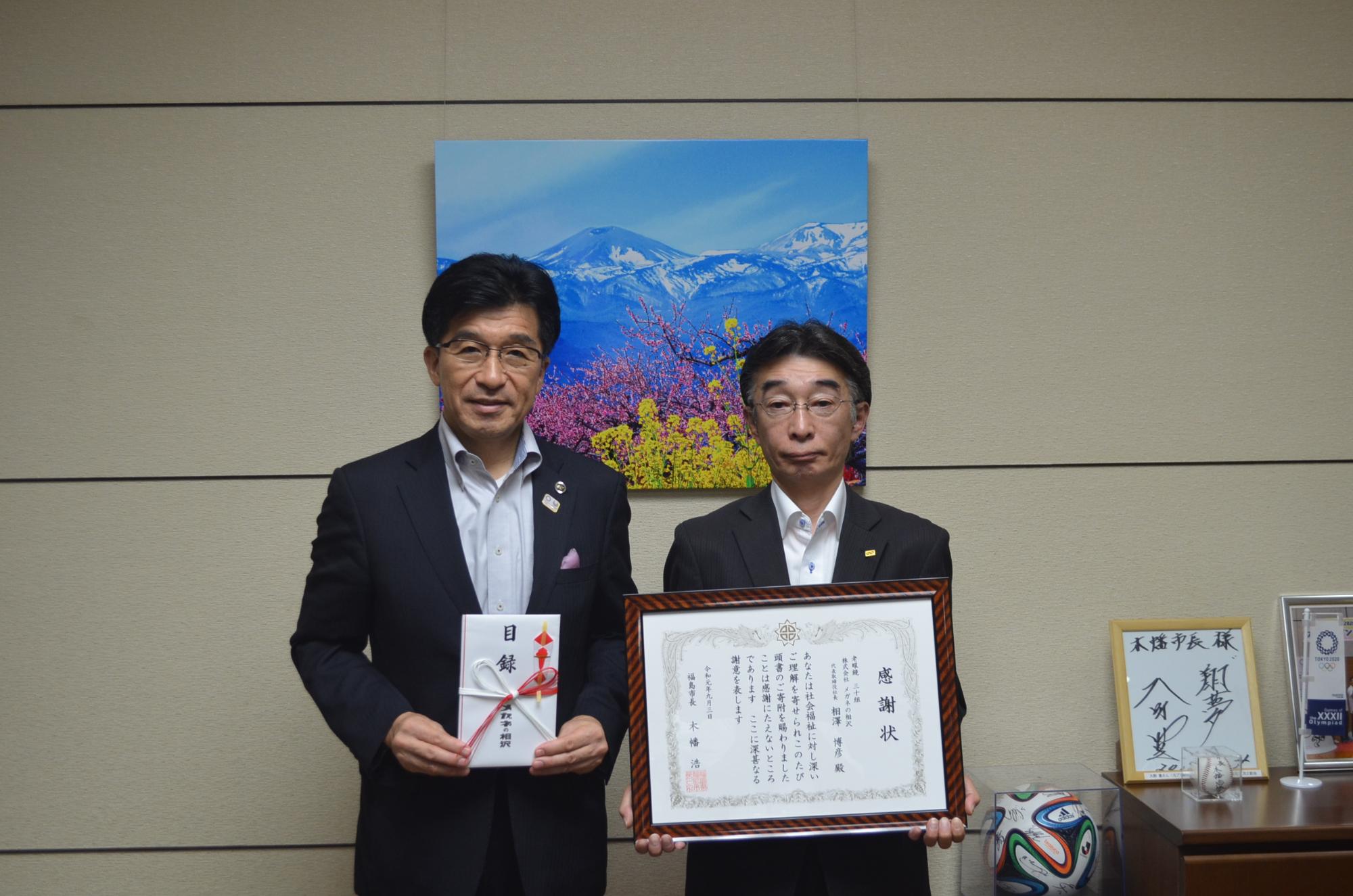 ピンク色と黄色の花と山の絵が飾られた室内でメガネの相沢の鈴木店長は額縁に入った感謝状を持ち、市長は金封を手に持って記念撮影している写真