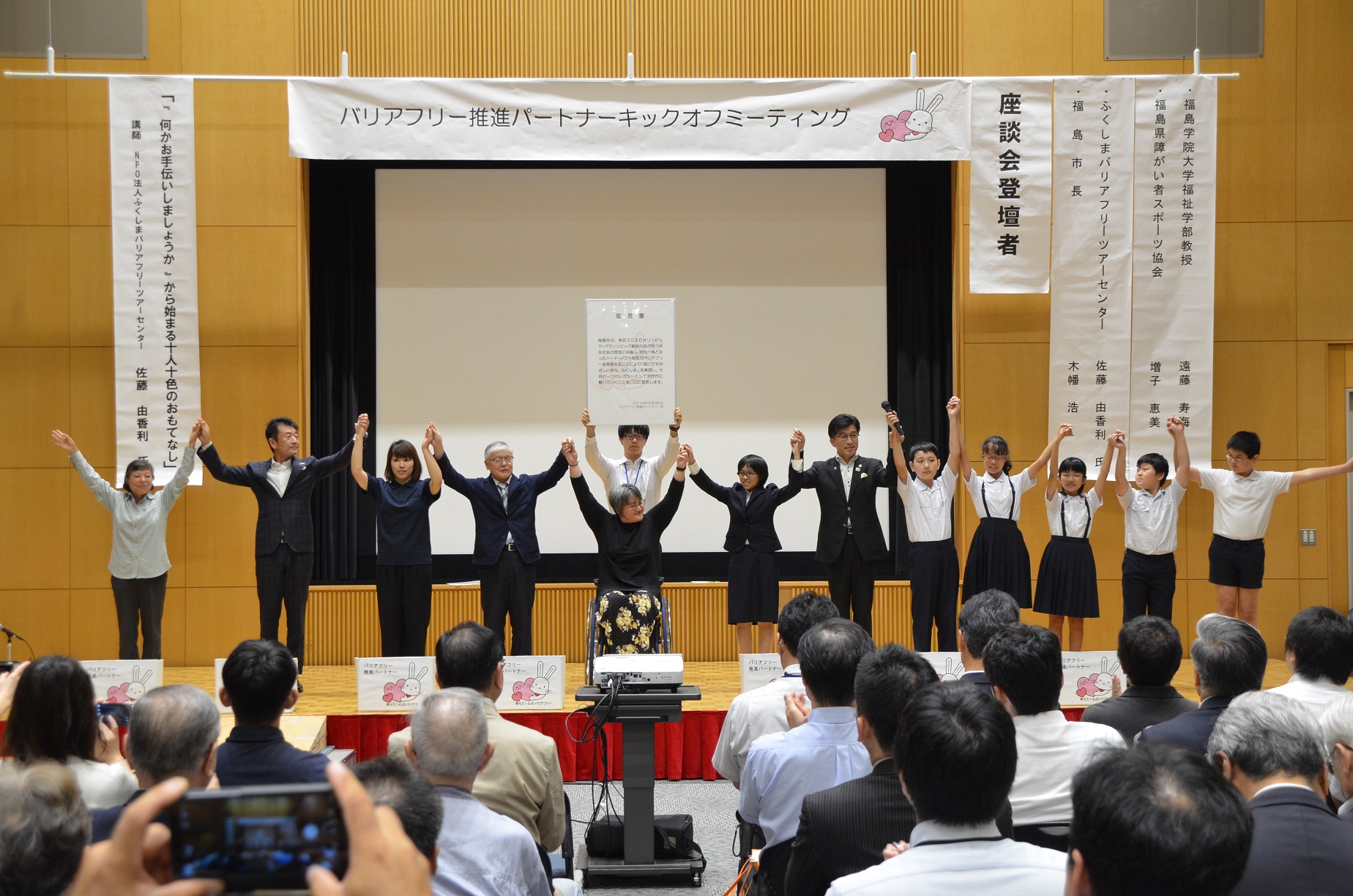 横断幕や垂れ幕が吊るされている下で、子供達や市長や大人の人達が横一列になって手を繋ぎ上にあげている写真