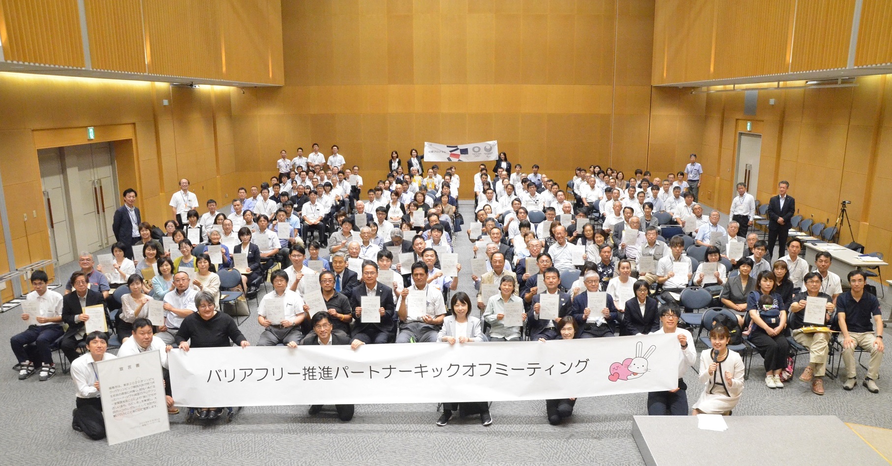 ホールの中で「バリアフリー推進パートナー キックオフミーテイング」と書かれた横断幕を持ち、整列された椅子に座った大勢の参加者達が並んでいる記念写真