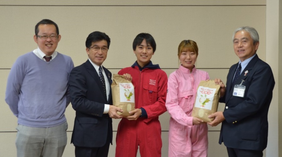 市長は赤色つなぎをきた女性と男性はピンク色のつなぎをきた女性とお米が入った米袋を一緒に持ち、市長の横にもう1人の男性が立ち、記念撮影している写真