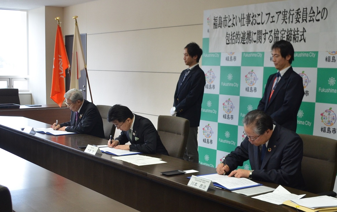「福島市とよい仕事おこしフェア実行員会との包括的連携に関する協定締結式」と書かれた横断幕が貼られた福島市のバックボードの前に男性2名が立ち、その前に設置された長机に市長と男性2名が座りサインをしている写真