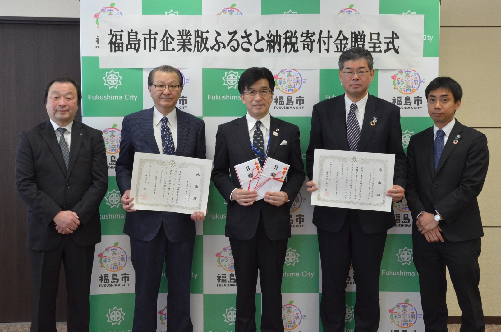 「福島市企業版ふるさと納税寄付金贈呈式」と書かれた横断幕が貼られた福島市のバックボードの前で金封を2つ手に持ち中央に立つ市長とスーツを着た男性4名が並び、市長の両隣りの男性2名は賞状もって記念撮影をしている写真
