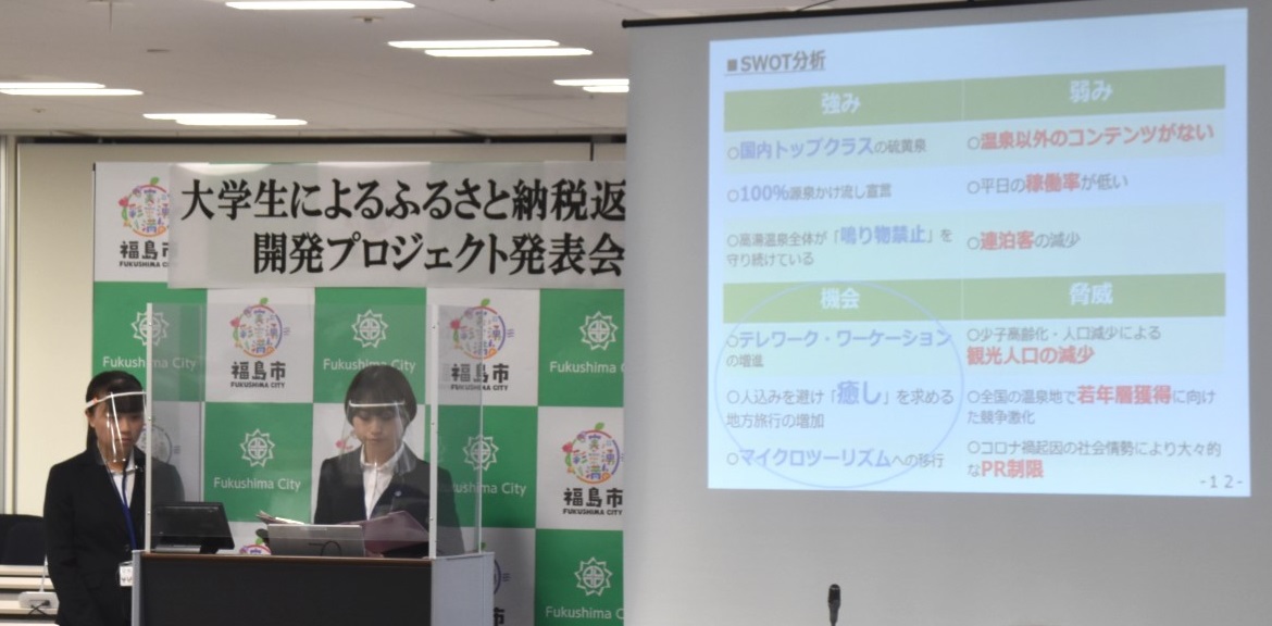 福島市のバックパネル前の演壇で、スクリーンに資料を投影しながら、発表をする女性の写真