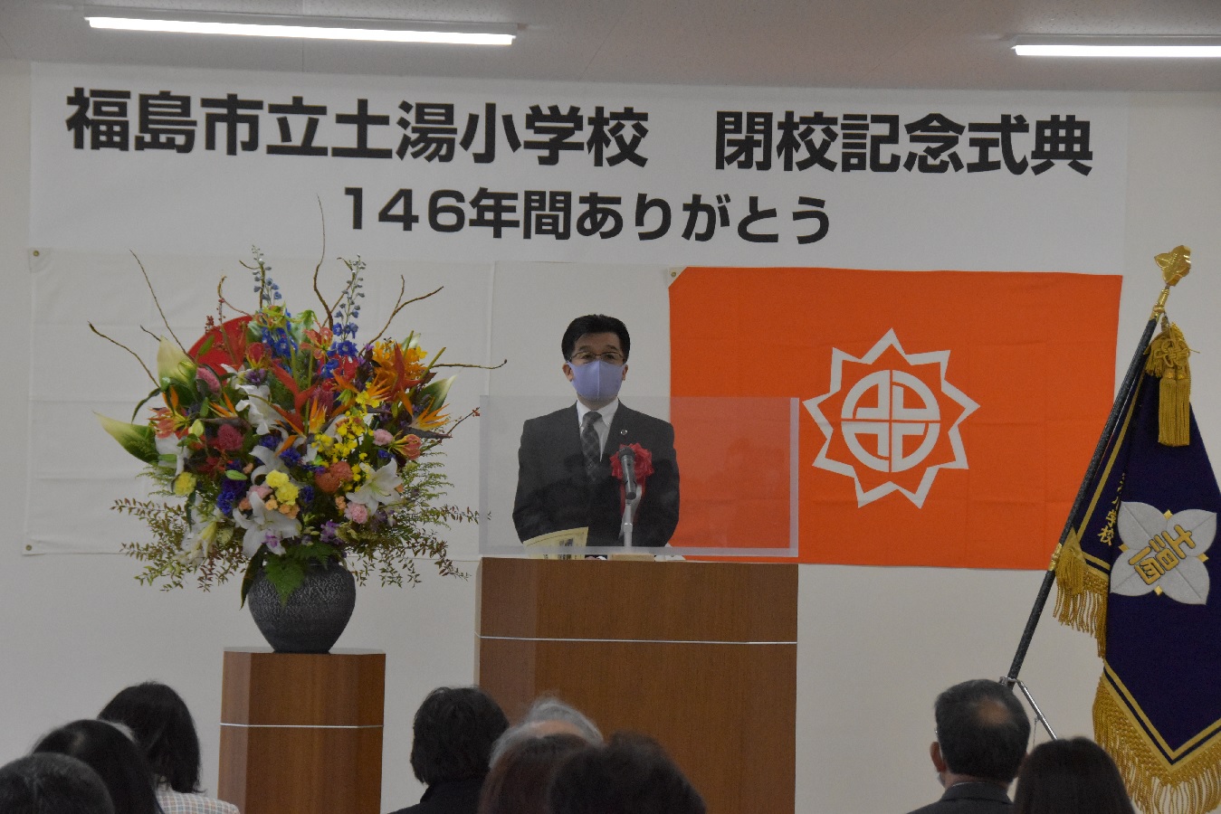 「福島市立土湯小学校 閉校記念式典 146年間ありがとう」の横断幕が掲げられ、両側に花と学校旗が飾られた演壇で話をする市長の写真