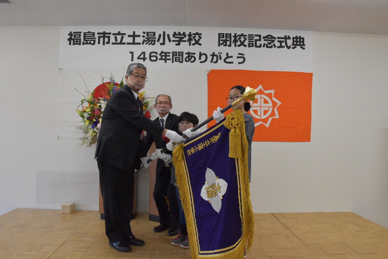 「福島市立土湯小学校 閉校記念式典 146年間ありがとう」の横断幕が掲げられた壁の前で、学校旗を手に持つ関係者と小学生達の写真