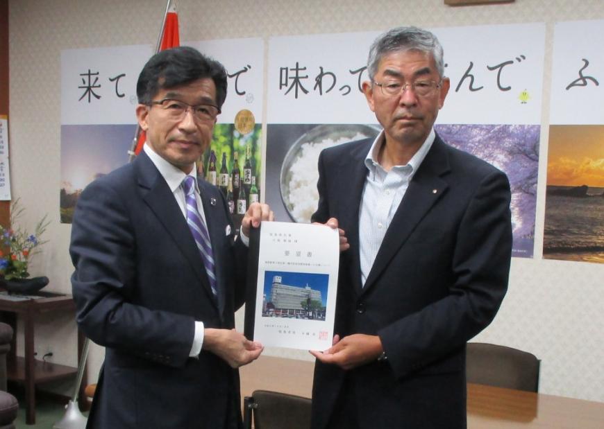 壁にポスターが貼られた執務室内で、市長と男性で要望書を持っている写真