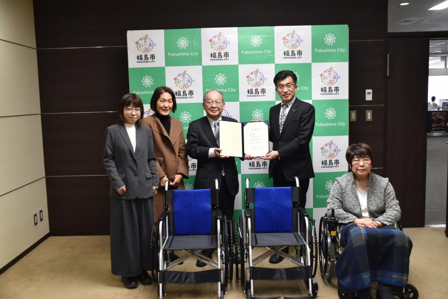 バックパネルの前に市長と男性で証書ファイルを持ち、横に女性2名が立ち、前に並べられた車いす3台の1台に女性が座っている写真