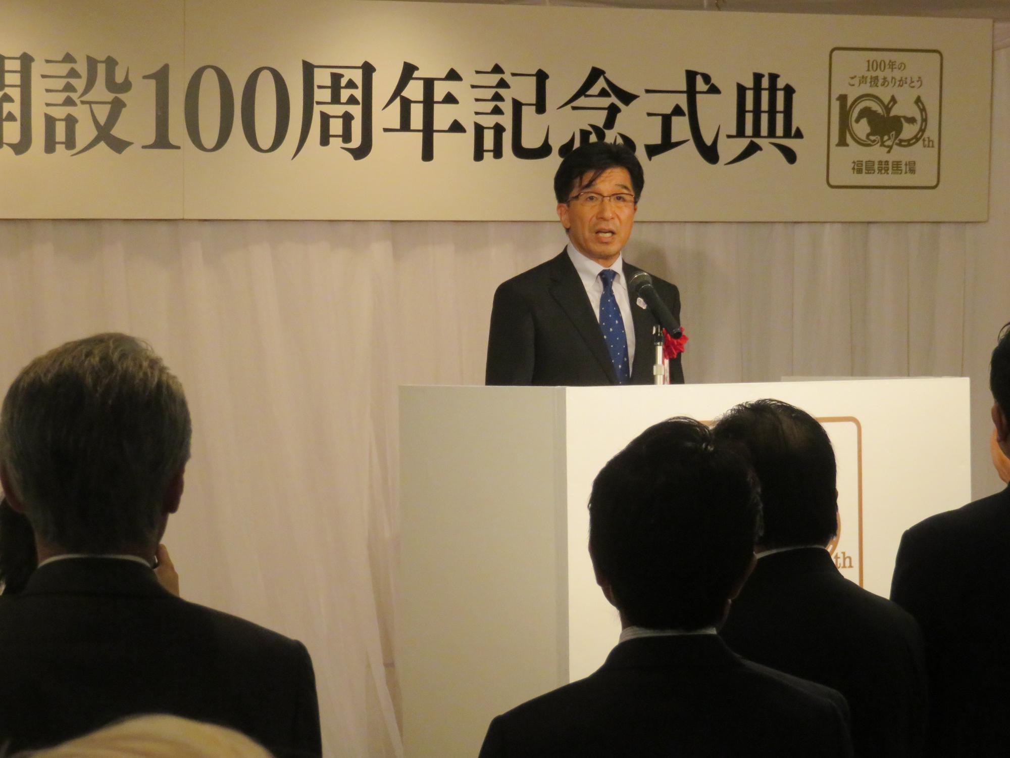 「福島競馬場開設100周年記念式典」のパネルが掛けられており、演壇で話をする市長の写真