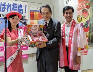 ピンクの帽子を被り、同色のワンピースを着た女性から桃が入った箱を手渡された男性の横に法被を着た市長が立っている写真