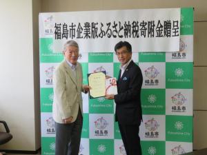 「福祉末子企業版ふるさと納税寄附金贈呈」と書かれた幕が貼ってあるバックパネルの前で、男性が賞状と目録を市長に手渡している写真