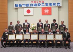 「福島市優良建設工事表彰式」と書かれた横断幕がある会場で、市長と関係者の間に額縁に入った賞状を持って7名の男性が座り、後方に胸章をつけた男性が7名立っている写真