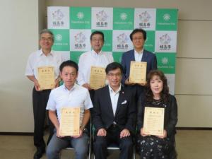 バックパネルの前で、市長の両脇に男性と女性が座り、その後ろに男性3名が立ち、それぞれ賞状を持っている写真