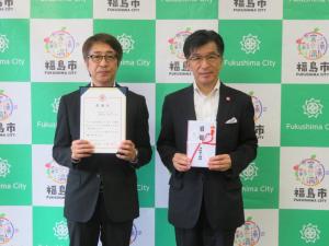バックパネルの前で、市長が目録、男性が賞状をそれぞれ持っている写真