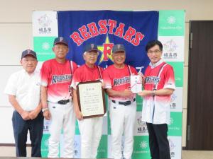 「REDSTARS」と書かれた幕が貼られたバックパネルの前で、ユニフォームの上着を着た市長とユニフォーム姿の3名の男性と半袖シャツを着た男性が立ち、賞状と目録をもっている写真