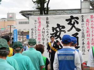 交通安全の横断幕がかけられた屋外の場所で、黄色の襷をかけた市長が青や緑の服を着て整列している参加者の前で話をしている写真