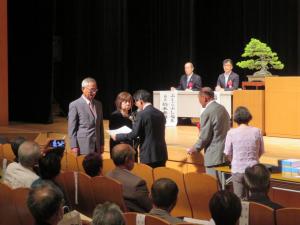 参加者が座っている前に立っている1組の夫婦に市長が賞状を読んでいる写真