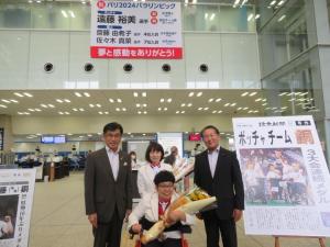 横断幕が掛けられ、ボッチャチームの新聞記事の看板がある会場で、花束を持ち車いすに乗った女性の後方に市長と男性1名、女性1名が立っている写真