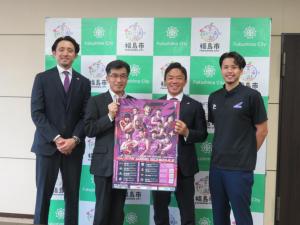 バックパネルの前で、市長と男性でポスターを持ち、その両脇に男性が1名ずつ立っている写真