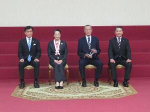 赤い絨毯の部屋で市長と男性の間にメダルをかけた女性と透明な盾を持った男性が座っている写真