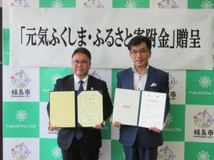 「元気ふくしま・ふるさと寄附金」贈呈と書かれた横断幕が貼られたバックパネルの前に立ち、市長と男性がお互いに証書ファイルをひろげている写真