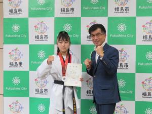 バックパネルの前でメダルを掛け賞状を持った道着姿の女の子と市長がガッツポーズをとっている写真