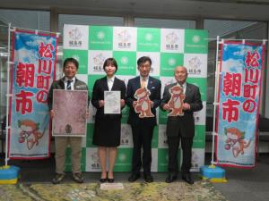 「松川町の朝市」と書かれた幟が立っている、バックパネルの前で、市長と男性がゆるキャラのまつカワウソくんのボードを、女性が任命書を、もう1名の男性がポスターをそれぞれ持って立っている写真