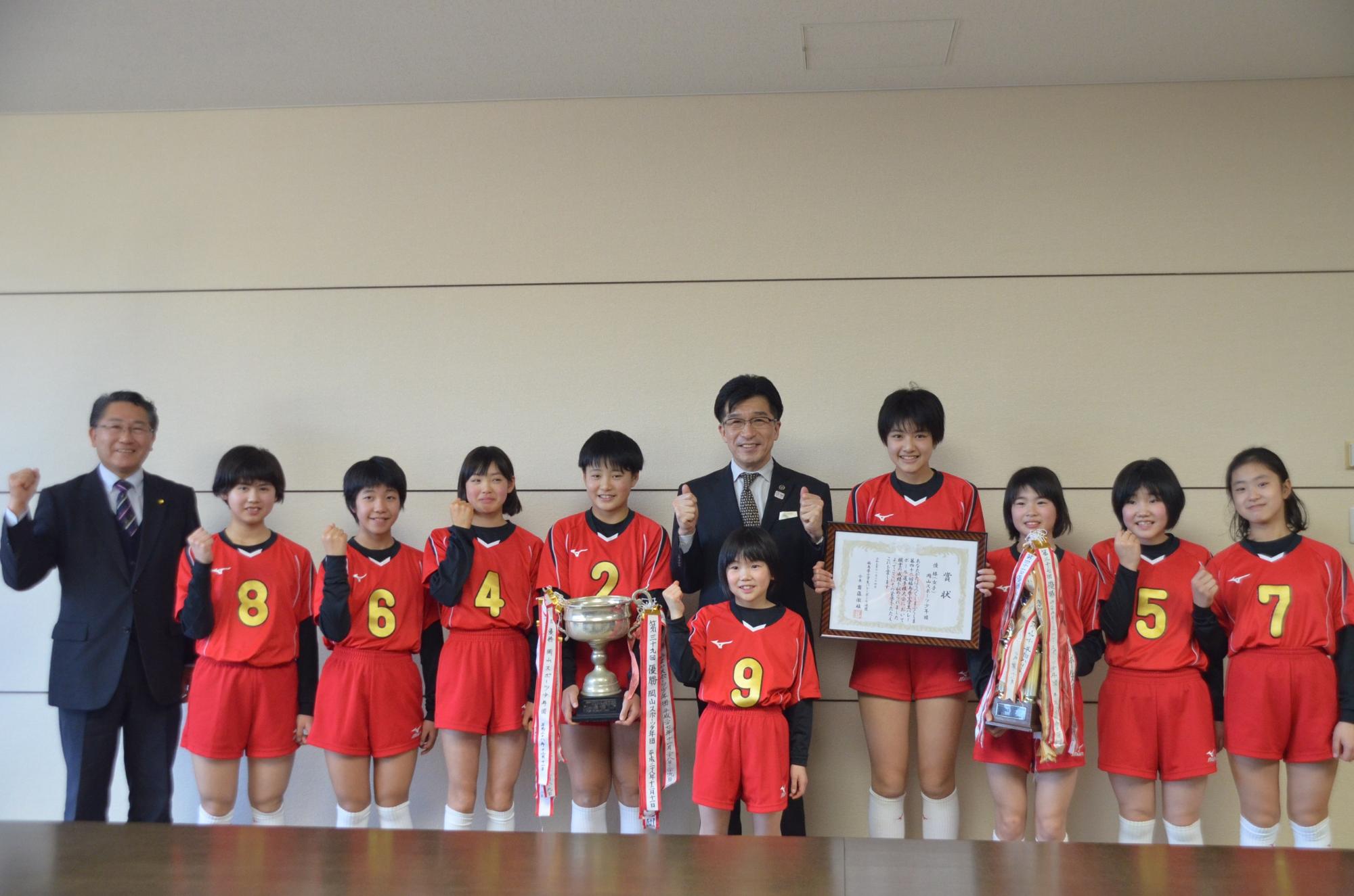 男性と赤いユニホームを着た岡山スポーツ少年団女子バレーボール部の皆さんと市長が横並びになり、ガッツポーズをして記念撮影をしている写真
