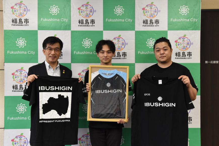 バックパネルの前で、市長と男性2名が[IBUSHIGIN」のロゴが入ったTシャツを持っている写真