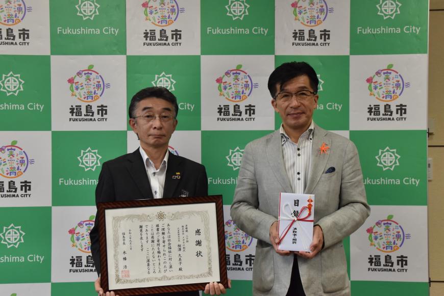 バックパネルの前で、市長が目録封筒を持ち、男性が額入りの感謝状を持って立っている写真