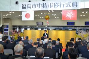 「福島市中核市移行式典」の横断幕と、国旗、福島市の旗が掲げられた会場の金屏風の前で、席に座った出席者たちに向かって話をしている市長の写真