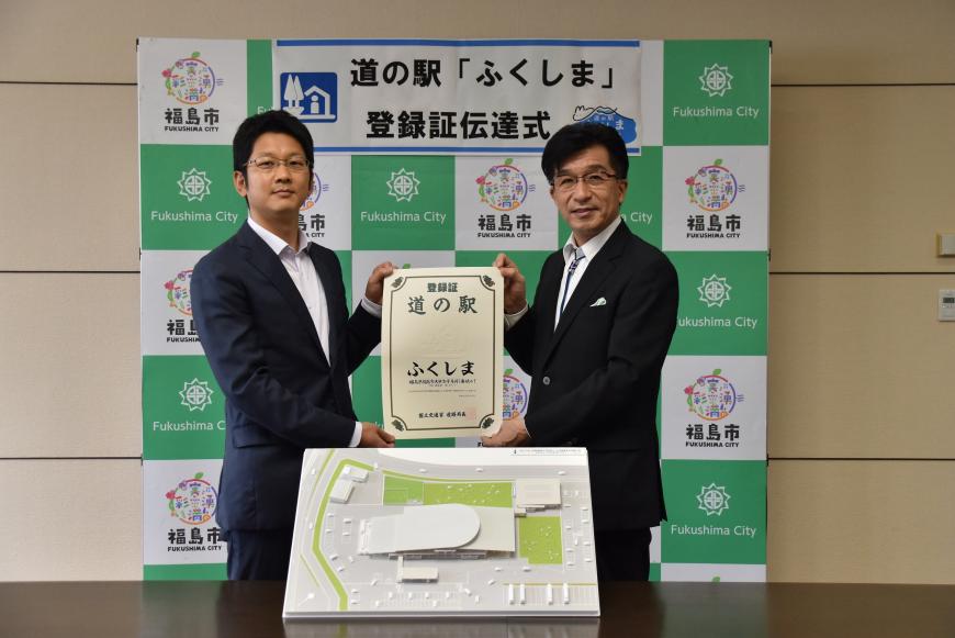 「道の駅ふくしま登録証伝達式と書かれた福島市のバックボードの前に立った市長とスーツの男性が、登録証を両側から持っており、その手前の机に道の駅の模型が置かれている写真