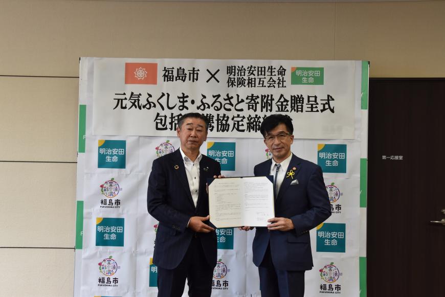 「元気ふくしま・ふるさと寄付金贈呈式包括連携協定」と書かれた横断幕が貼られた明治安田生命と福島市のロゴマークが格子状に並べられたバックボードの前で市長と男性が協定書を一緒に持ち、記念撮影している写真